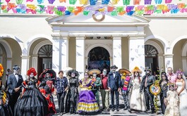 Aparecen personas vestidos como catrines y catrinas en edificio con adornos de papel de china de colores festejando el dia.