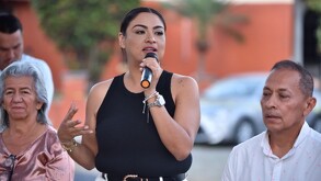 Fotografía en plano medio de una mujer con cabello oscuro recogido, vestida con una camiseta sin mangas negra y jeans blancos, sosteniendo un micrófono negro.