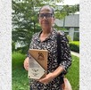 Una mujer de mediana edad con lentes y blusa estampada sostiene un reconocimiento de madera por donación de órganos, flanqueada por el logo del IMSS y un símbol Una mujer de mediana edad con lentes y blusa estampada sostiene un reconocimiento de madera por donación de órganos, flanqueada por el logo del IMSS y un símbol