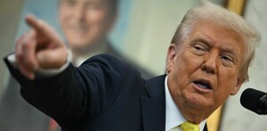 Donald Trump, con traje oscuro y corbata amarilla, habla en un atril mientras señala enfáticamente hacia la izquierda. Un retrato sonriente está borroso al fond