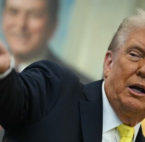 Donald Trump, con traje oscuro y corbata amarilla, habla en un atril mientras señala enfáticamente hacia la izquierda. Un retrato sonriente está borroso al fond Donald Trump, con traje oscuro y corbata amarilla, habla en un atril mientras señala enfáticamente hacia la izquierda. Un retrato sonriente está borroso al fond