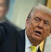 Donald Trump, con traje oscuro y corbata amarilla, habla en un atril mientras señala enfáticamente hacia la izquierda. Un retrato sonriente está borroso al fond