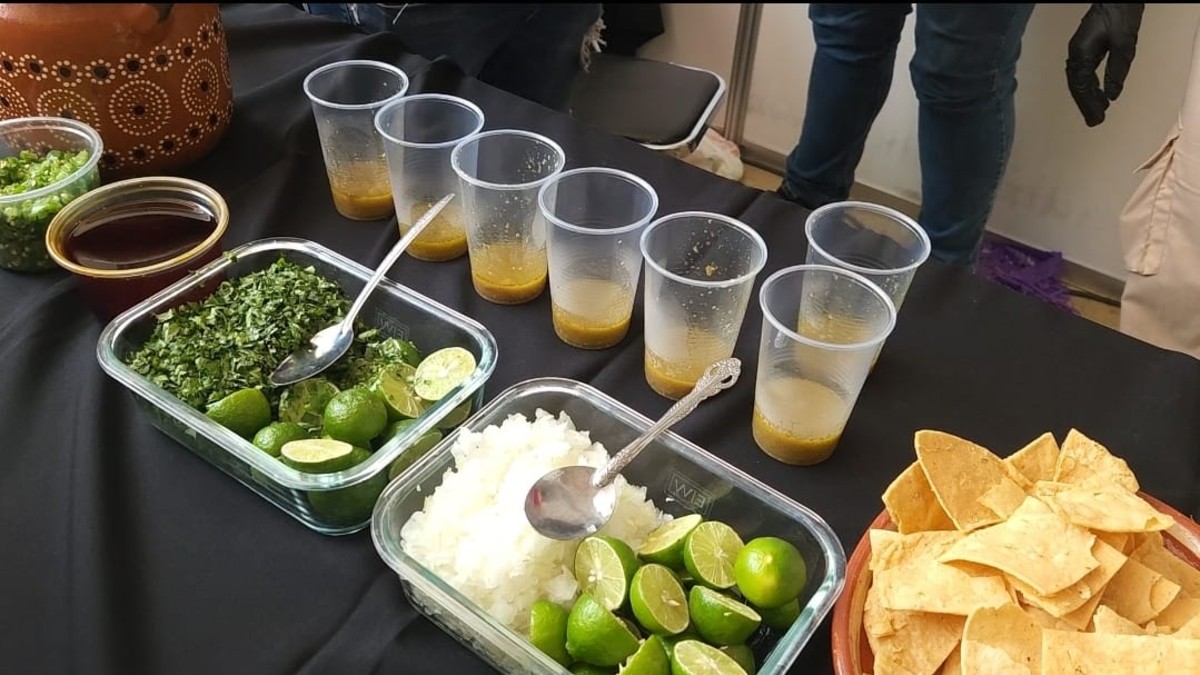 Mesa con ingredientes de carne en su jugo: limones, cilantro, cebolla picada y totopos, junto a varios vasos con el caldo del guiso. Fondo negro y persona de pi Mesa con ingredientes de carne en su jugo: limones, cilantro, cebolla picada y totopos, junto a varios vasos con el caldo del guiso. Fondo negro y persona de pi