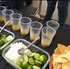 Mesa con ingredientes de carne en su jugo: limones, cilantro, cebolla picada y totopos, junto a varios vasos con el caldo del guiso. Fondo negro y persona de pi