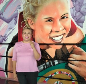 Una mujer en playera rosa posa frente a un mural de boxeo vibrante que muestra a una campeona sonriente con un cinturón. Se aprecian las cuerdas y esquinas Una mujer en playera rosa posa frente a un mural de boxeo vibrante que muestra a una campeona sonriente con un cinturón. Se aprecian las cuerdas y esquinas