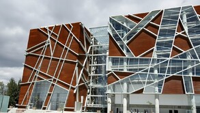 Edificio moderno con fachada de madera marrón y estructura geométrica de vidrio y metal blanco bajo un cielo nublado y gris.