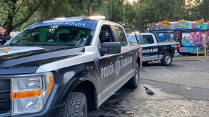 Primer plano de una camioneta de la Policía de Guadalajara estacionada en la calle, con otra patrulla detrás. Al fondo se distingue un área de juegos mecánicos