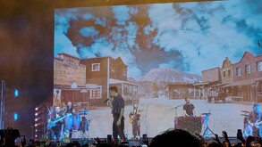 Pepe Madero cantando en un escenario con una gran pantalla de fondo que muestra un pueblo del viejo oeste y montañas. Su banda toca a los lados.