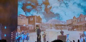 Pepe Madero cantando en un escenario con una gran pantalla de fondo que muestra un pueblo del viejo oeste y montañas. Su banda toca a los lados. Pepe Madero cantando en un escenario con una gran pantalla de fondo que muestra un pueblo del viejo oeste y montañas. Su banda toca a los lados.