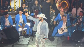 El cantante Pancho Barraza en el escenario con un traje de mezclilla brillante y sombrero vaquero, levanta la mano. Está rodeado por su banda de mariachi