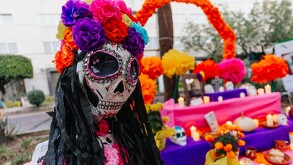 Primer plano de una figura de La Catrina con un cráneo decorado y una corona de flores de papel brillantes (rosa, morado, azul). Un altar del Día de Muertos