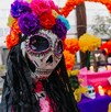 Primer plano de una figura de La Catrina con un cráneo decorado y una corona de flores de papel brillantes (rosa, morado, azul). Un altar del Día de Muertos