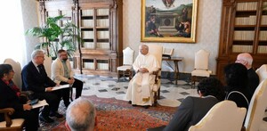 León XIV sentado en el centro, con vestimenta blanca, preside una reunión con varias personas en un salón con libreros de madera y un cuadro.