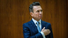 Retrato del político Pablo Lemus con traje azul y corbata clara, con la mano sobre el pecho. Mira hacia su derecha con expresión seria. Fondo de pared de madera