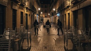 Tres hombres encapuchados corren por una calle empedrada de noche, cada uno cargando varias sillas metálicas, mientras dos patrullas de policía