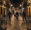 Tres hombres encapuchados corren por una calle empedrada de noche, cada uno cargando varias sillas metálicas, mientras dos patrullas de policía Tres hombres encapuchados corren por una calle empedrada de noche, cada uno cargando varias sillas metálicas, mientras dos patrullas de policía