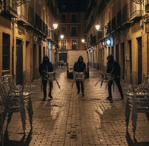 Tres hombres encapuchados corren por una calle empedrada de noche, cada uno cargando varias sillas metálicas, mientras dos patrullas de policía