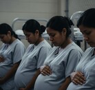 Cuatro mujeres embarazadas migrantes con uniformes grises idénticos, sentadas en una hilera tocando sus vientres. El fondo sombrío sugiere una detención