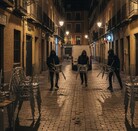 Tres hombres encapuchados corren por una calle empedrada de noche, cada uno cargando varias sillas metálicas, mientras dos patrullas de policía