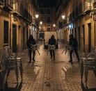 Tres hombres encapuchados corren por una calle empedrada de noche, cada uno cargando varias sillas metálicas, mientras dos patrullas de policía
