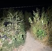 Camino de tierra angosto y oscuro por la noche, flanqueado por maleza y arbustos altos. Un área de basura y escombros se distingue a la izquierda, iluminada por Camino de tierra angosto y oscuro por la noche, flanqueado por maleza y arbustos altos. Un área de basura y escombros se distingue a la izquierda, iluminada por