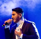 En la imagen aparece Joan Sebastián Alarcón, quien se encuentra con un atuendo en color negro sosteniendo un micrófono, en lo que parece ser un concierto.