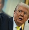 Donald Trump, con traje oscuro y corbata amarilla, habla en un atril mientras señala enfáticamente hacia la izquierda. Un retrato sonriente está borroso al fond Donald Trump, con traje oscuro y corbata amarilla, habla en un atril mientras señala enfáticamente hacia la izquierda. Un retrato sonriente está borroso al fond