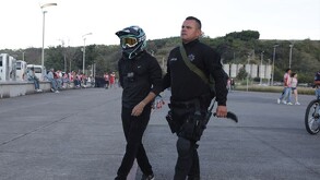 Un oficial de policía con uniforme oscuro y equipo táctico camina con un detenido esposado que lleva un casco de motocross. Al fondo se ve a público de fútbol