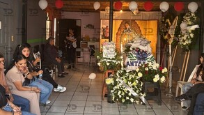 Un funeral con un ataúd cubierto y arreglos florales rodeados por una ofrenda de la Virgen de Guadalupe y globos. Varias personas, principalmente mujeres.
