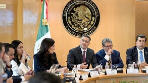 El funcionario Omar García Harfuch habla en una mesa de trabajo en el Congreso, gesticulando con la mano. Detrás de él está el político Ricardo Monreal escuchan