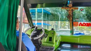 Chofer manejando una unidad del transporte público en color verde y con una cruz de la religión católica.