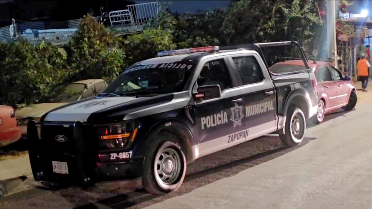 Vista lateral de noche de una patrulla de la Policía de Zapopan frente a unas plantas y con automóviles alrededor Vista lateral de noche de una patrulla de la Policía de Zapopan frente a unas plantas y con automóviles alrededor