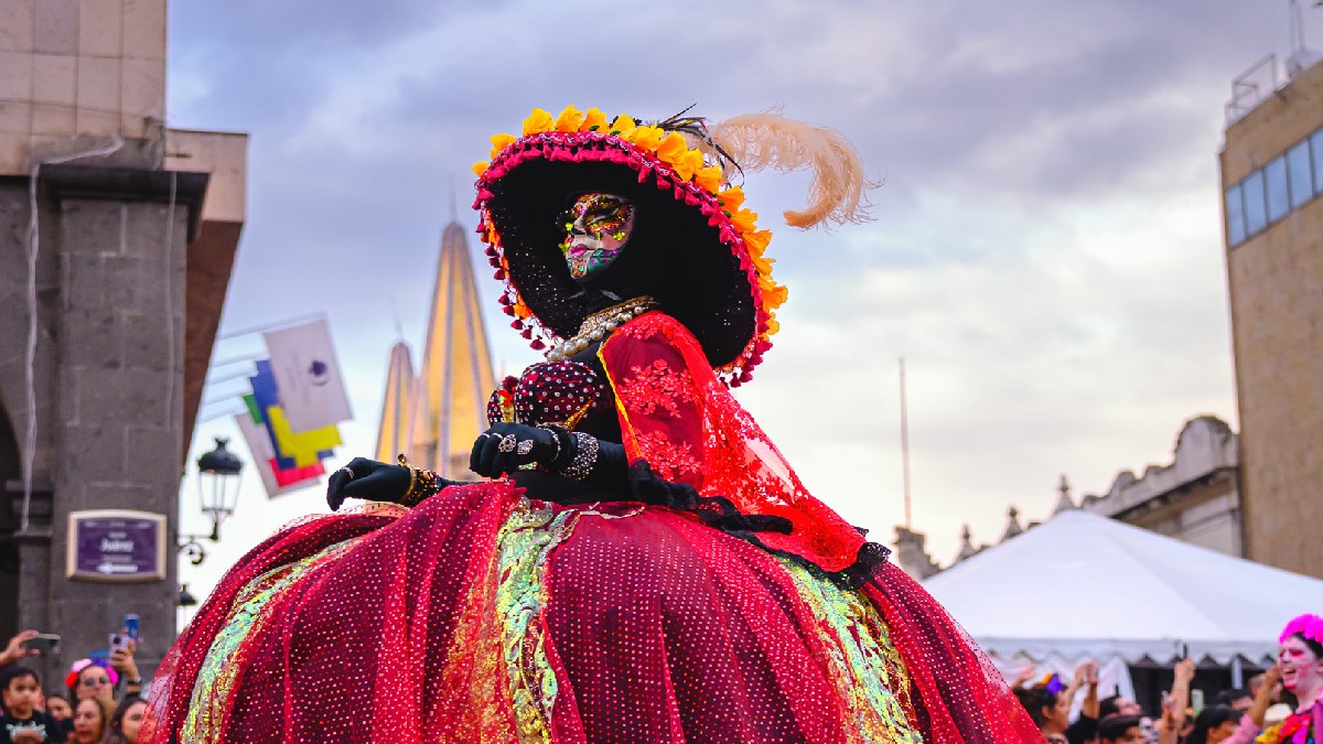Una mujer vestida de Catrina con un enorme y colorido vestido rojo y un sombrero grande con flores y plumas. Posa al aire libre en el centro de la ciudad