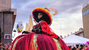 Una mujer vestida de Catrina con un enorme y colorido vestido rojo y un sombrero grande con flores y plumas. Posa al aire libre en el centro de la ciudad