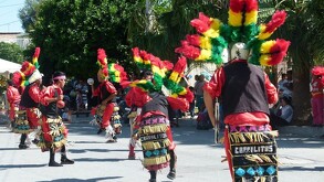 En la imagen se puede ver un gurpo de danzantes con vestimenta en color rojo y penacho color verde y amarillo.