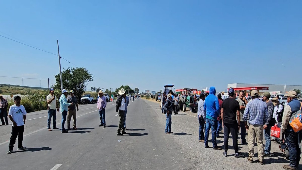 Una toma de plano medio muestra un grupo de hombres, varios usando sombreros de ala ancha, parados sobre una carretera asfaltada que ha sido bloqueada.