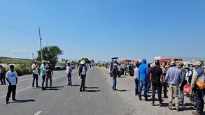 Una toma de plano medio muestra un grupo de hombres, varios usando sombreros de ala ancha, parados sobre una carretera asfaltada que ha sido bloqueada.