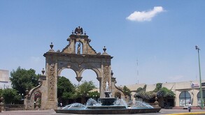 Vista diurna de Los Arcos de Guadalajara con su diseño barroco. En primer plano, una fuente con agua y chorros, y en el fondo, edificios modernos y la calle