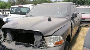 Una camioneta pick-up negra, de doble cabina, abandonada o detenida en un terreno baldío. La puerta del conductor está abierta. Hay hierba seca y un autobús