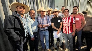 Grupo de campesinos y ciudadanos se reúnen de noche frente a una reja, muchos usando sombreros vaqueros. Uno de ellos viste la camiseta de fútbol de Chivas