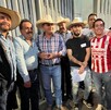 Grupo de campesinos y ciudadanos se reúnen de noche frente a una reja, muchos usando sombreros vaqueros. Uno de ellos viste la camiseta de fútbol de Chivas