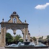 Vista diurna de Los Arcos de Guadalajara con su diseño barroco. En primer plano, una fuente con agua y chorros, y en el fondo, edificios modernos y la calle