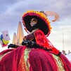 Una mujer vestida de Catrina con un enorme y colorido vestido rojo y un sombrero grande con flores y plumas. Posa al aire libre en el centro de la ciudad Una mujer vestida de Catrina con un enorme y colorido vestido rojo y un sombrero grande con flores y plumas. Posa al aire libre en el centro de la ciudad