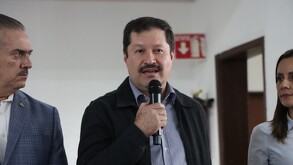 El director del Mercado de Abastos de Guadalajara habla por micrófono en un evento. Viste una chaqueta oscura y camisa a cuadros. A su lado, un hombre y una muj