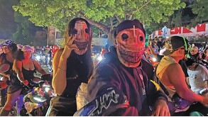 Primer plano de una pareja en motocicleta durante una rodada nocturna. Ambos usan máscaras de terror con ojos de costura. Están rodeados por otros motociclistas