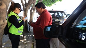 Mujer con chaleco fosforescente frente a un hombre de camisa roja hablan junto a un vehículo estacionado.