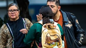 Un hombre da un beso en la frente a su hija antes de dejarla en la escuela; junto a él, una señora de la tercera edad.