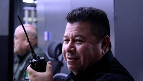 Primer plano de un policía jubilado sonriente. Viste una camisa negra y sostiene un radio comunicador. En el fondo, desenfocado, hay otro colega. El ambiente es