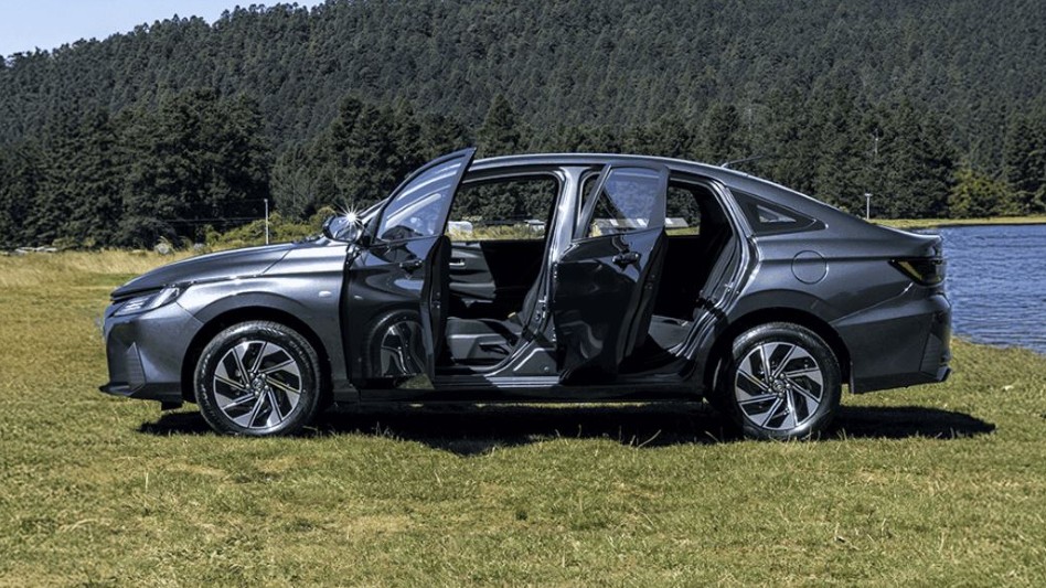 Vista lateral del Toyota Yaris Sedán HEV 2026 color gris oscuro con las puertas abiertas sobre el pasto en un día soleado y detrás un lago y un bosque de pinos