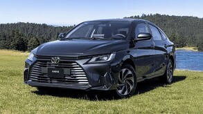 Vista frontal del Toyota Yaris Sedán HEV 2026 color fgris oscuro sobre el pasto en un día soleado y detrás un lago y un bosque de pinos